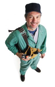 a caucasian male plummer in a green jumpsuit and a ballcap holds his tools and looks up at the camera